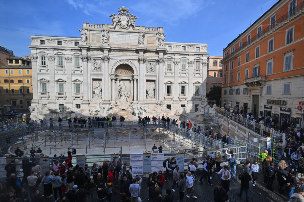 Panoramic view of the Trevi Fountain being renovated with tourists throwing coins into a temporary pool from an elevated walkway in Rome.